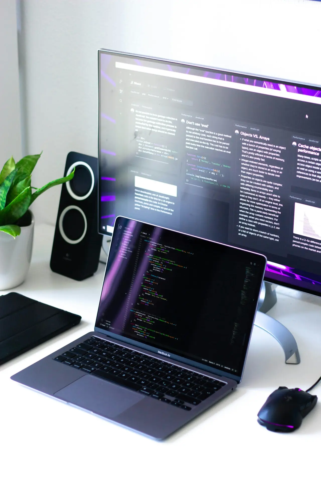 MacBook Air menampilkan kode di layar, diletakkan di atas meja putih bersih dengan monitor eksternal, speaker, mouse gaming RGB, dan tanaman hias.
